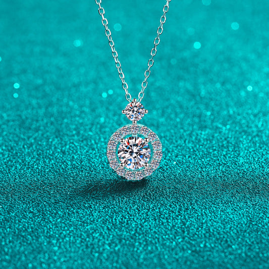 One carat Moissanite necklace set in 925 Sterling silver with a large stone in the center, smaller stone at the base of the chain, and small stones surrounding the center stone, on a teal surface.