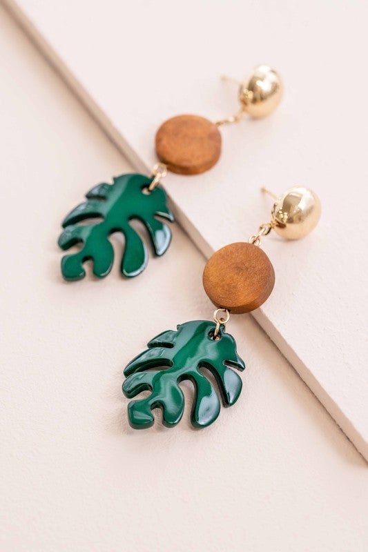 Tropical palm leaf dangle earrings with gold accents and a wooden disc on a flat surface.