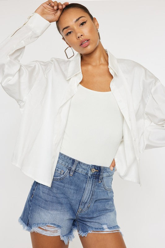 Model with dark hair wearing a long sleeve white shirt over a white tank with the high rise denim mom shorts, distressed.