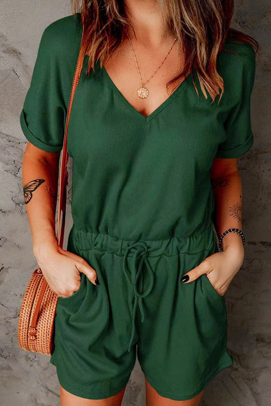 Brunette model wearing a kelly green drawstring waist, v-neck, rolled short sleeve romper against a concrete wall. Available in many colors.