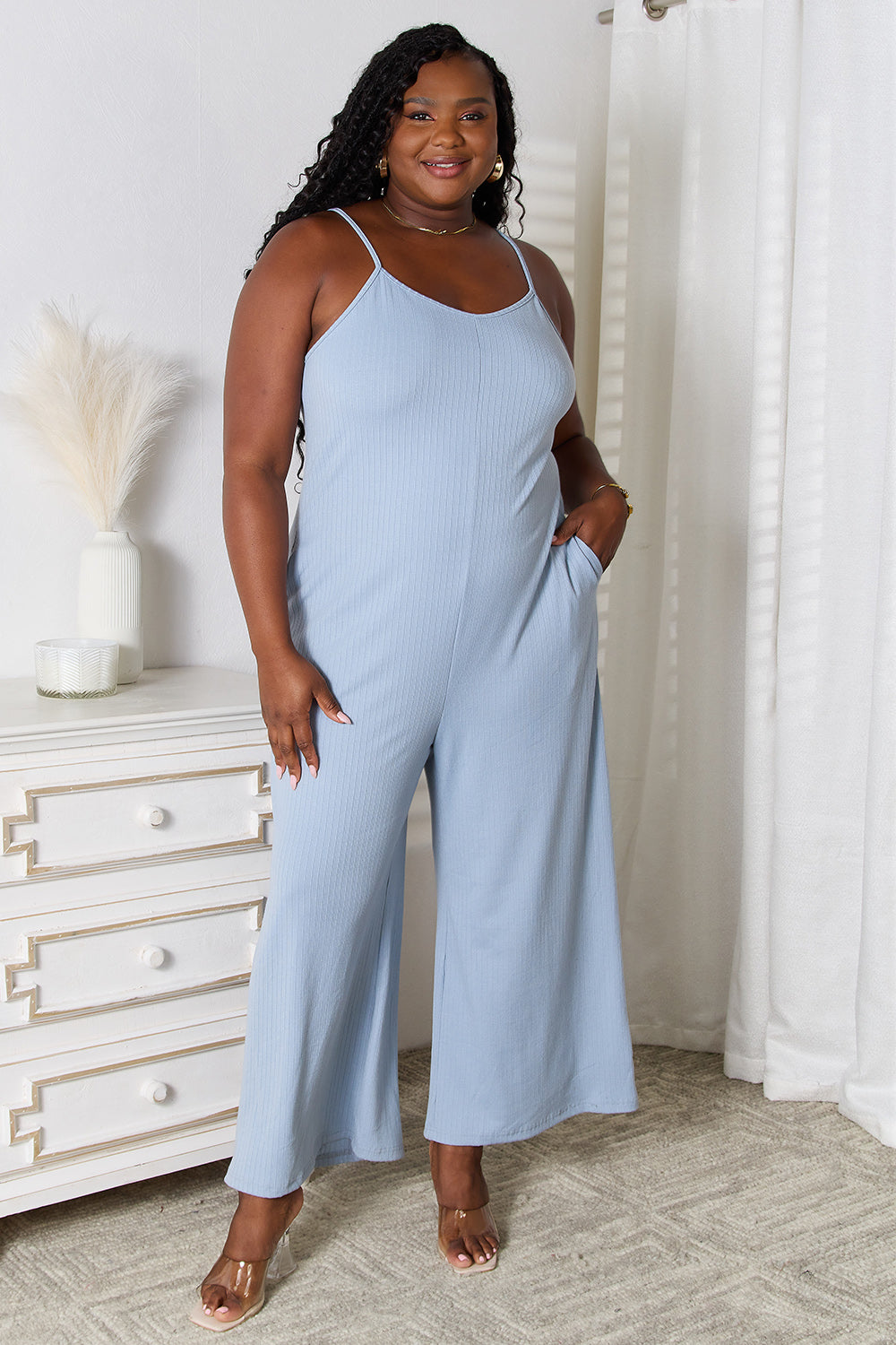 Dark-skinned model with long dark hair wearing a light blue jumpsuit with pockets, spaghetti straps, and wide legs against a white bedroom background.