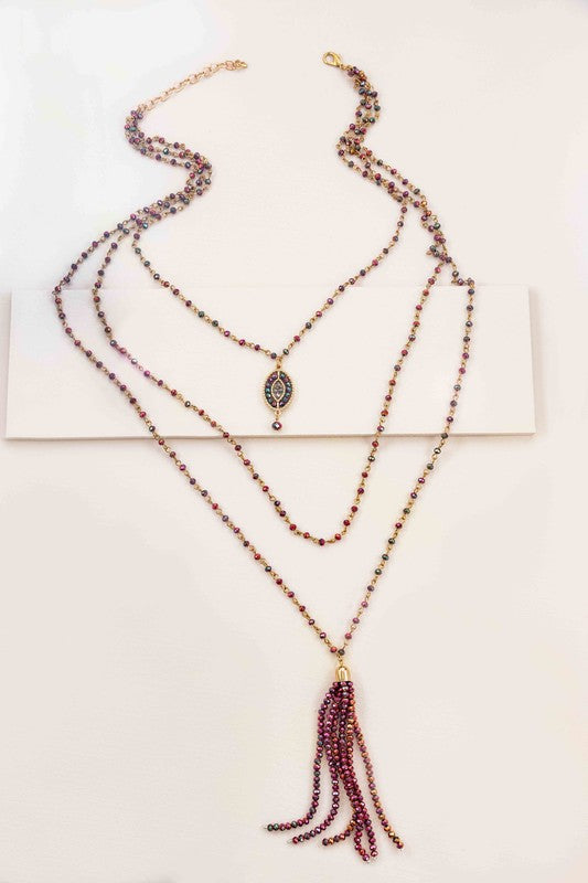 Three layer purple and green beaded necklace with a short strand with a small medaliion, a medium strand of all beads, and a long strand with a beaded tassel at the end, on an off-white background.