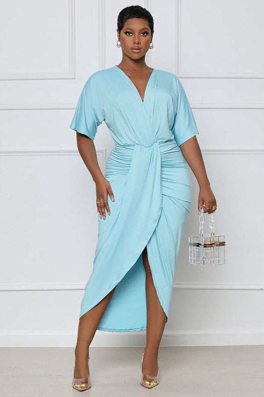 Beautiful sky blue midi dress with a petal bottom, ruched waist, short sleeves, and a surplice top worn by a brunette model against a white wall.