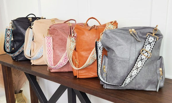 Convertible vegan leather backpack/tote bags in black with teal strap, tan with light blue strap, pink with pink strap, Orange with orange strap, and grey with black strap on a wooden table.