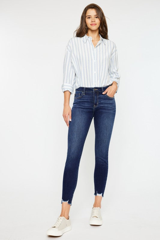 Brunette model wearing a blue and white vertical striped long sleeved shirt with the KanCan mid rise Ankle length skinny jeans with frayed hem.