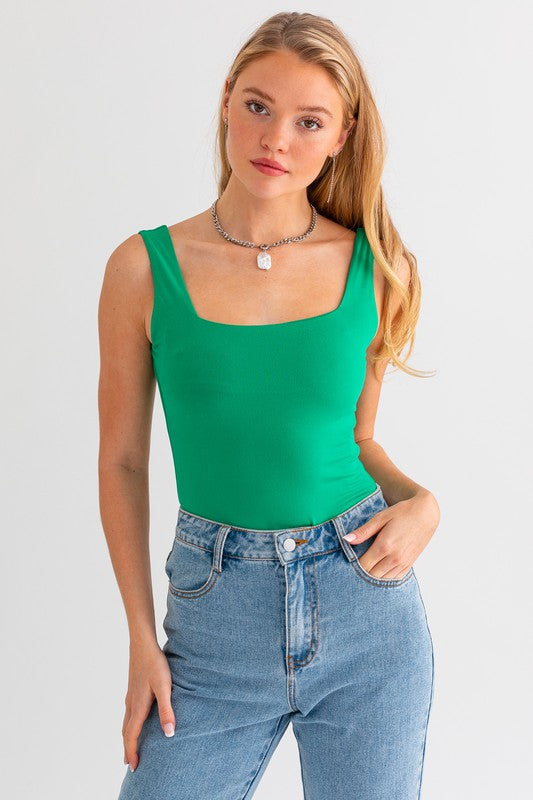 Blonde model wearing jeans with a square neck tank bodysuit in kelly green against a white wall.