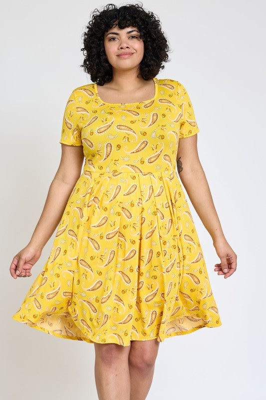 Brunette model wearing a flirty and fun yellow paisley midi dress with short sleeves, a full skirt, and a square neckline against a white backdrop.