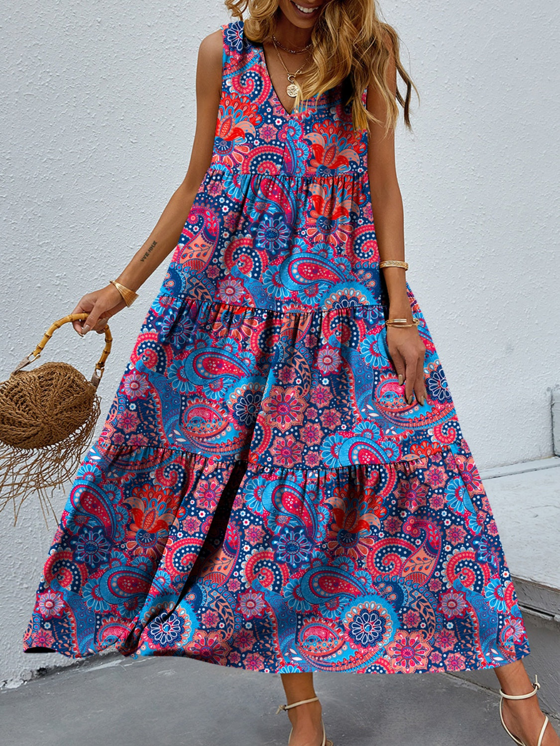 Bright paisley and floral print tiered, printed, v-neck, sleeveless casual dress worn by a blonde model against a concrete wall.