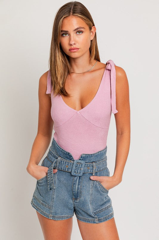 Pink sleeveless bodysuit with tie straps and a deep v-neck on a blonde model wearing jean shorts against a white backdrop.