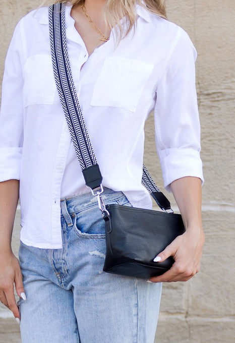 Blonde model wearing a white shirt and light blue jeans accessorized by a black genuine leather compact crossbody bag with a guitar strap against a concrete wall.