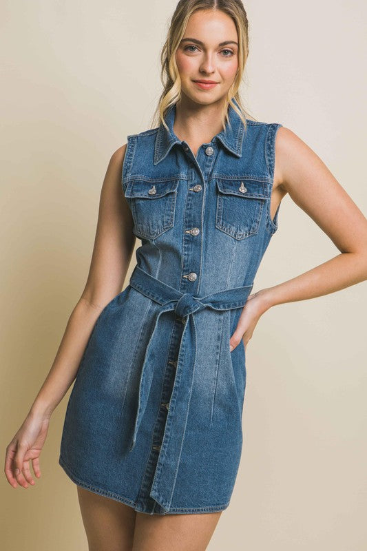 Blonde model wearing a medium blue denim mini dress with a tie waist, breast pockets, and collar in front of an ecru backdrop.