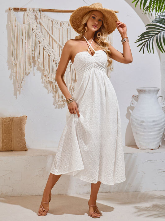 Blonde model in a white living room wearing a white Swiss dot, halter neck midi sundress with sandals and a straw hat.