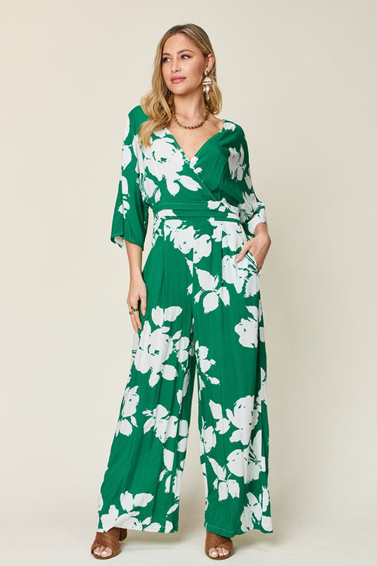 Blonde model wearing a half sleeve tropical printed tie back wide leg jumpsuit in kelly green with white flowers against an ecru background.