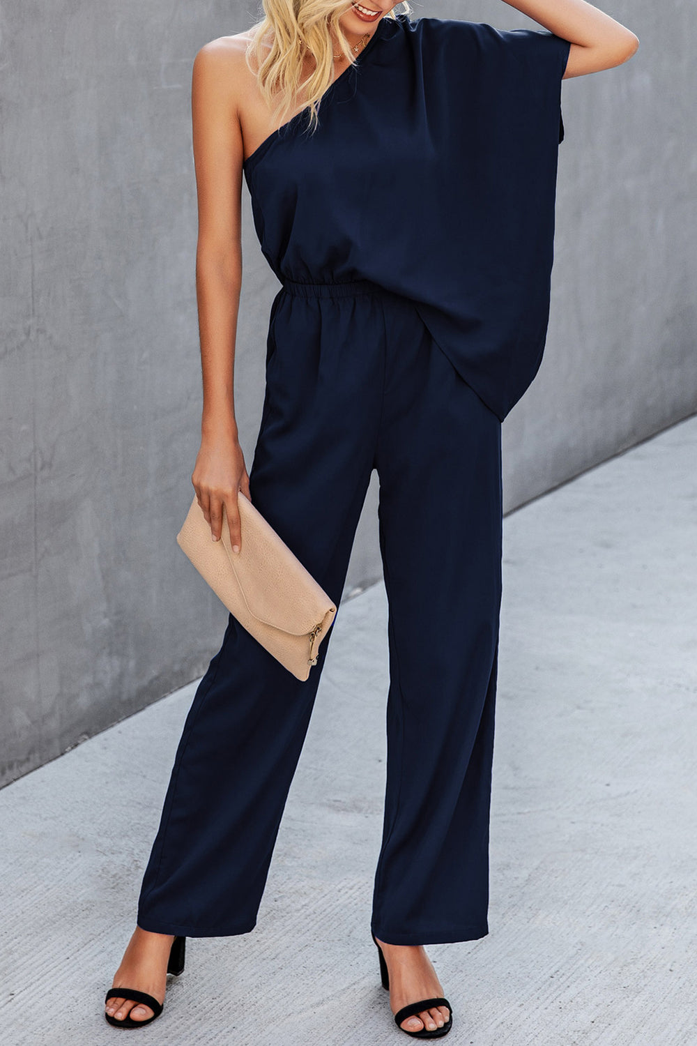 One shoulder jumpsuit in navy worn by a blonde model in front of a concrete wall.