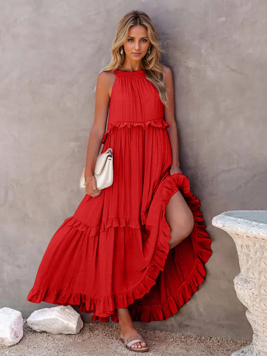 Blonde model leaning against a concrete wall wearing a red sleeveless tiered maxi dress with ruffle accents on tiers. 