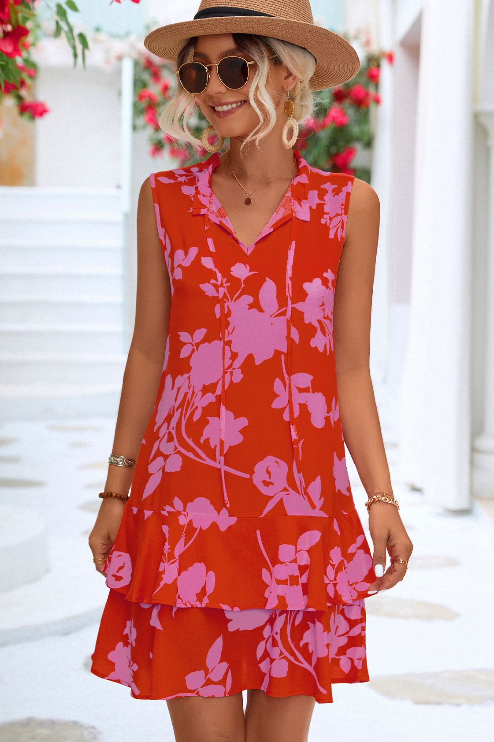 Blonde model in tropical setting wearing a printed tie neck sleeveless  tiered dress in a red and pink tropical print