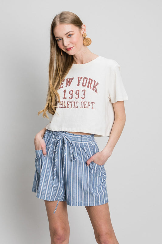Young blonde model wearing a white and red New York tee with a pair of Cotton Blue by Nu Label yarn dyed blue and white striped shorts with pockets, and a drawstring elastic waist.