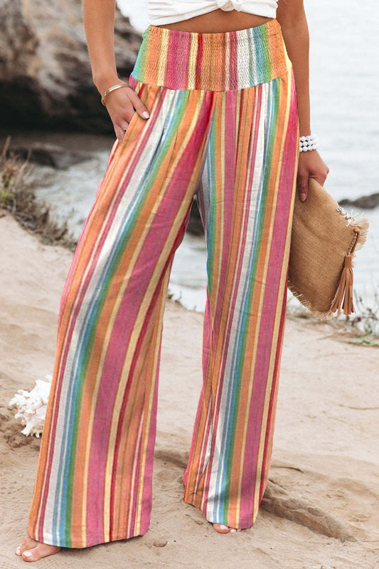 Model on a beach wearing a pair of smocked waist, wide leg, multicolor stripe pants with pockets.