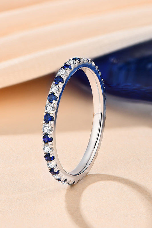 Slender eternity ring turned on it's side with alternating stones of lab-grown sapphires and moissanite. Shown on a peach and blue background