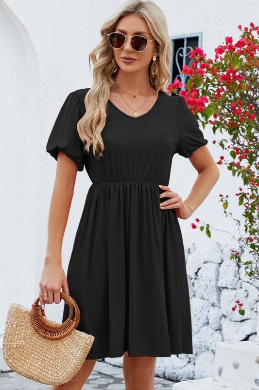 Blonde model in front of a flowering tree wearing a black, v-neck, short balloon sleeve dress that falls above the knee with an elastic waist.