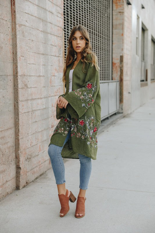 Brunette model wearing an olive green kimono with colorful embroidered flowers, made of lightweight gauzy polyester with a bell sleeve on a city street.