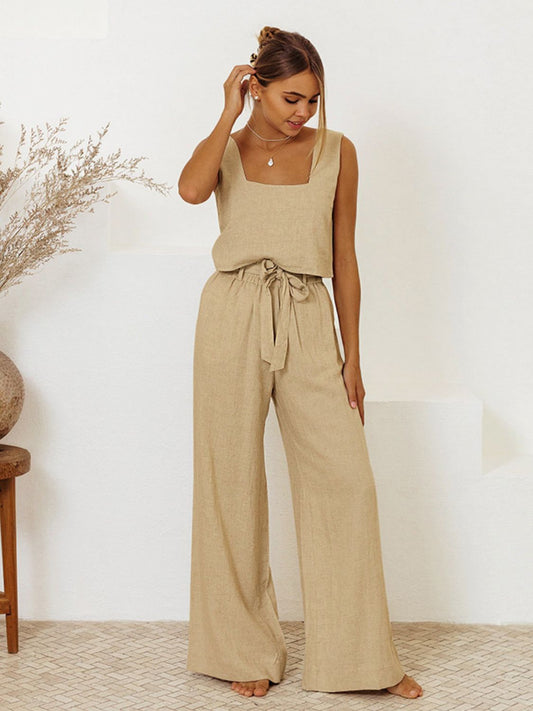 Brunette model in front of a white wall wearing a tan top and bottom set with wide leg, tied waist pants and a square neck, sleeveless top for a cool and comfortable summer.