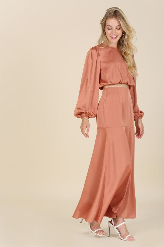 Blonde model wearing a round neck, long balloon sleeve satin top in brick with a matching satin maxi skirt bottom. Wear together or as separates.