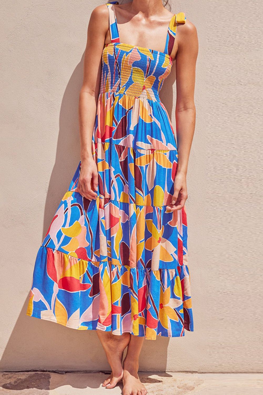 Model in front of a stucco wall wearing a multicolor, bright midi dress with a large floral print, tie shoulders, smocked top, and tiered bottom.