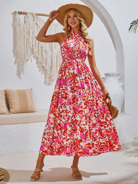 Blonde model in a white living room wearing a smocked, tiered, strawberry floral midi sundress with a multiway tied neck with sandals and a sun hat.