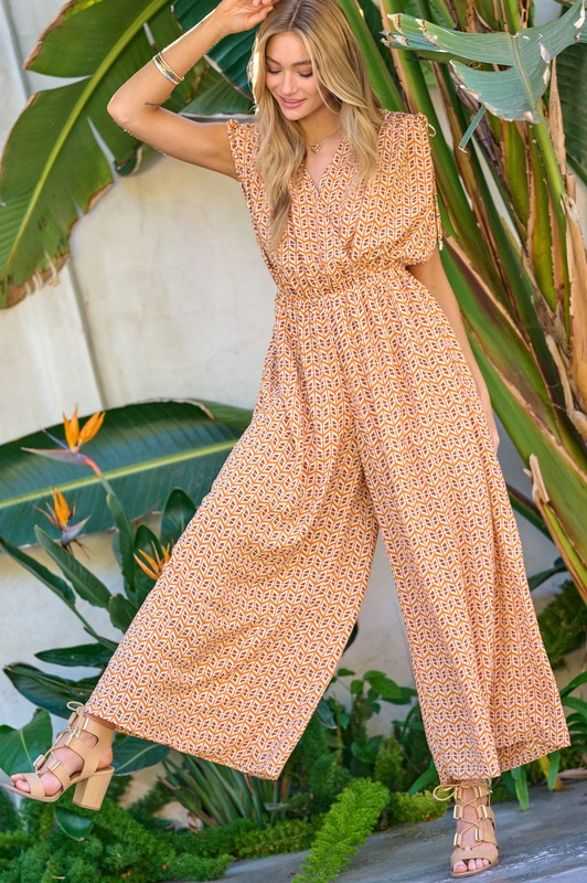 Blonde model wearing an orange printed v-neck sleeveless jumpsuit with tan heels against a greenery background.