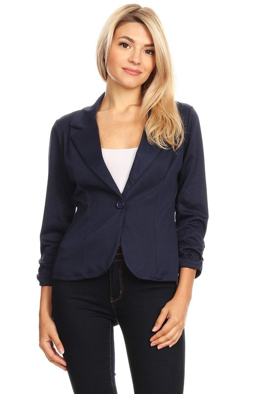 Blonde model wearing a Moa Collection waist length button front ruched sleeve blazer in navy against a white wall.