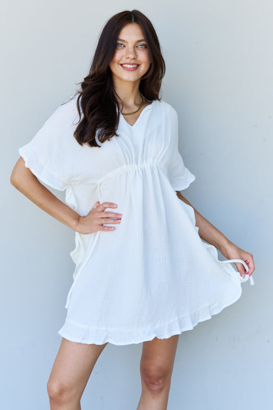 Brunette model wearing a 100% cotton white mini dress with a v-neck, ruffle details, and a drawstring waistband.
