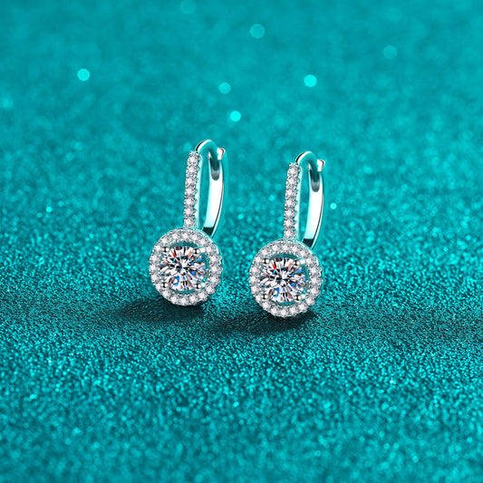 Pair of Moissanite drop earrings set in sterling silver with half-carat center moissanite surrounded by smaller stones on a teal surface.