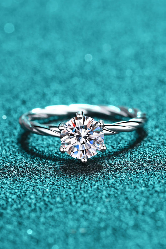 One carat moissanite solitaire ring set in rhodium plated sterling silver with a brushed surface twisted band. Shown on a teal. background.