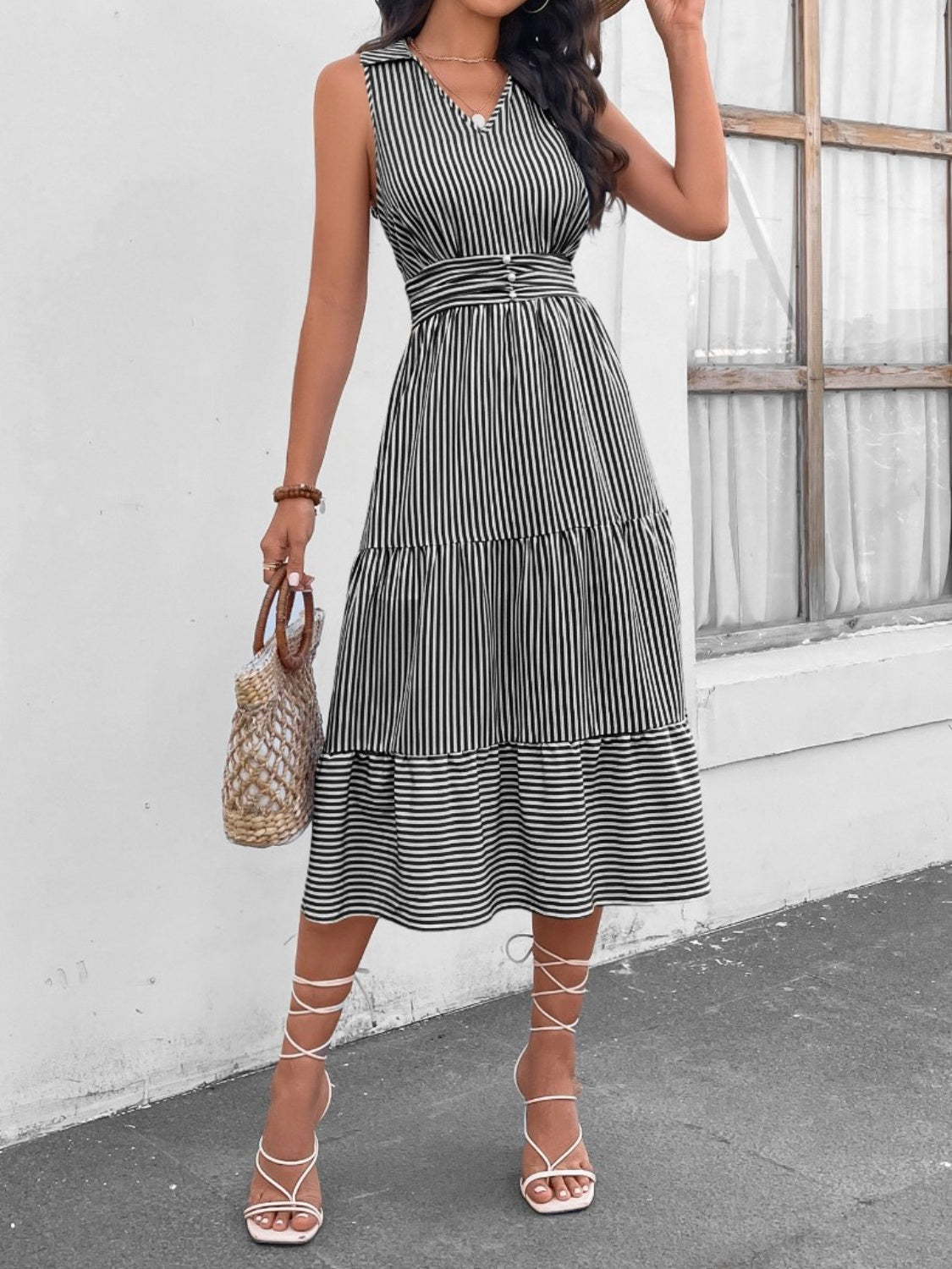 Brunette model in front of a white wall and window wearing a striped Johnny collar sleeveless midi dress with a tiered hem in thin black and white stripes. Available in multiple colors.