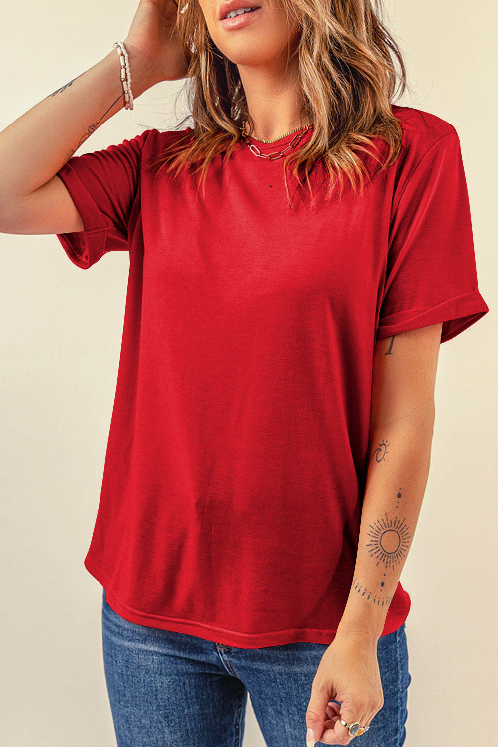 Model with dirty blonde hair wearing jeans and showcasing a red round neck, short sleeve tee with rolled sleeves against a white background.