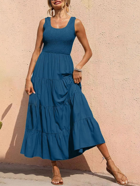 Blonde model in front of a peach wall wearing a peacock blue sleeveless smocked top tiered hem midi dress.