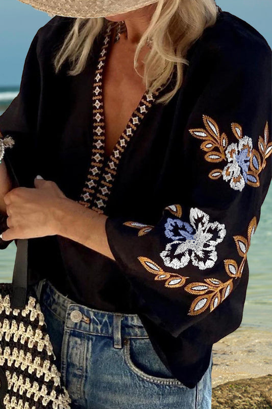 Blonde model in blue jeans on a beach wearing a black 3/4 sleeve 100% cotton blouse with white, light blue, and tan embroidery.