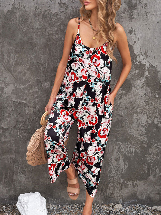 Blonde model wearing a black, red, and turquoise floral printed jumpsuit with spaghetti straps and pockets against a concrete wall.