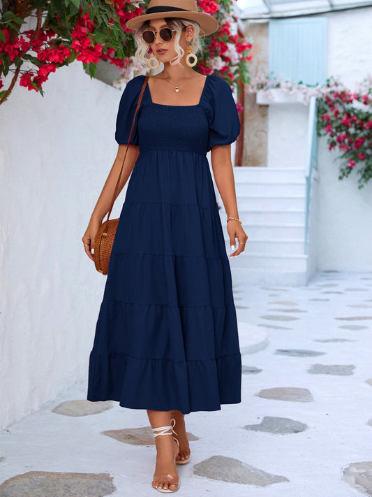 Blonde model in front of red flowers wearing a tiered, smocked short sleeve dress in navy blue.