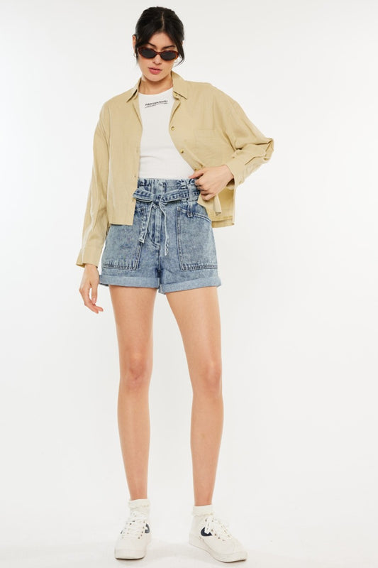 Brunette model wearing sunglasses with a white tank, khaki overshirt, and the KanCan ultra high waist paperbag denim shorts in medium denim color against a white backdrop.