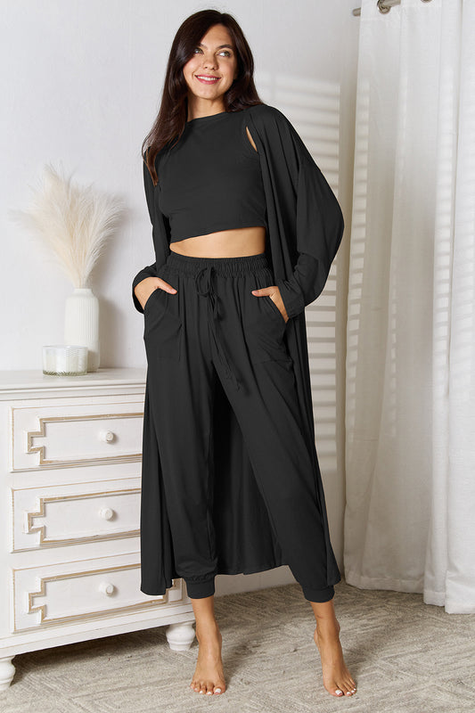 Brunette model in front of a white dresser wearing a black cropped tank, elastic waist jogger pants, and duster length cardigan set. Three pieces, one price.
