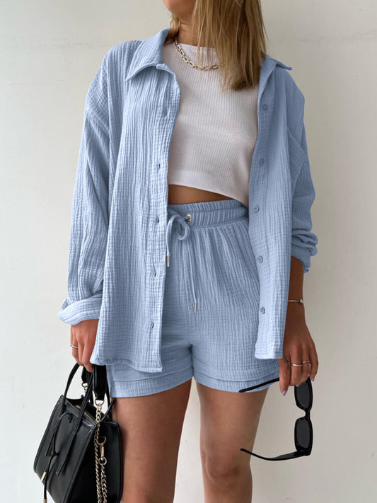 Blonde model wearing a white crop top with a two-piece textured button up long sleeve shirt and drawstring shorts set in light blue. Available in 12 colors.
