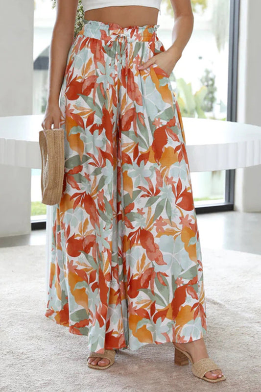 Model in a dining room wearing a pair of tropical print wide leg pants with pockets and a tied waist.