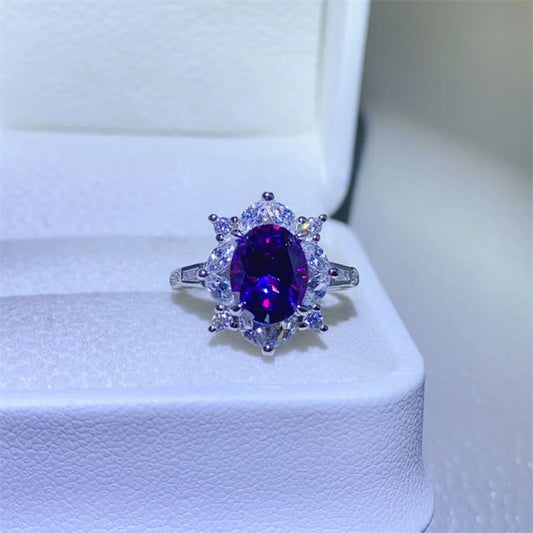Beautiful 2 carat Moissanite ring set in sterling silver with a large oval deep purple center stone surrounded by clear moissanite smaller stones. Photographed on top of a white ring box with a white background.