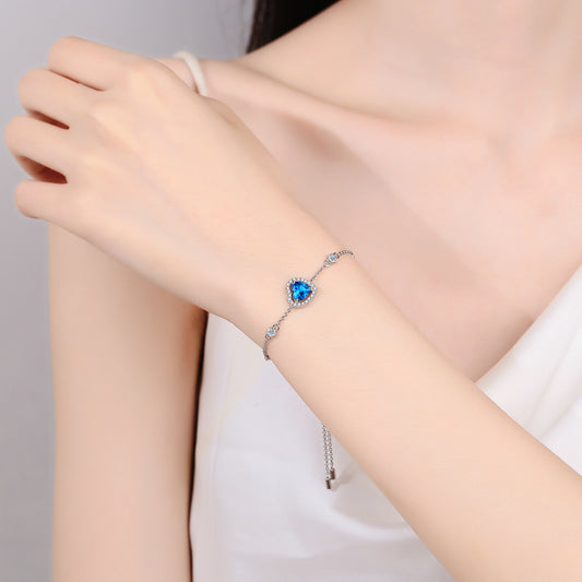 Model with dark hair wearing a white shirt wears a ultra marine blue heart shaped Moissanite bracelet. Blue heart in the center surrounded by smaller clear stones, with two more stones in the chain to each side of the heart.